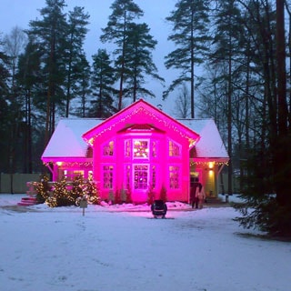 На фото: вечер, одноэтажный коттедж в скандинавском стиле, с праздничной подсветкой розового цвета, на участке - выпавший снег, на заднем плане - корабельные сосны